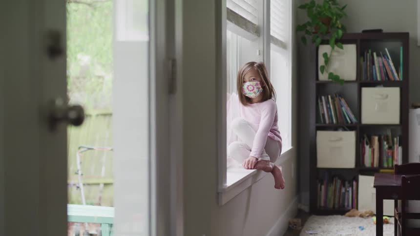 A child is looking out the window while wearing a face mask