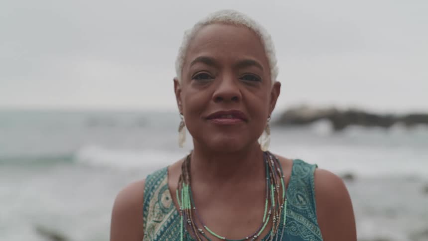 Black woman with earrings and short grey hair standing on the beach by the sea smiles and looks at the camera on a cloudy day
