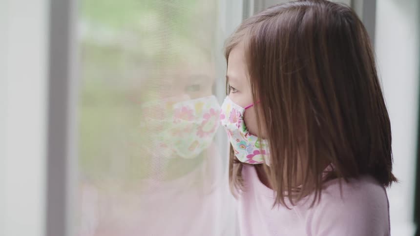Asian little girl with pink long sleeve t shirt and a face mask looks out the window