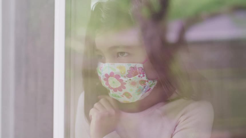 Asian little girl with a face mask looks out the window feeling bored stiff