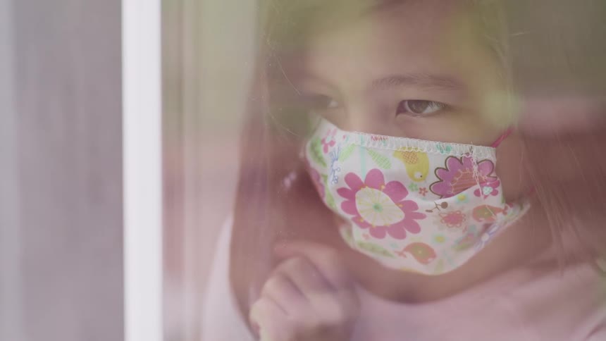 Asian little girl with brown straight hair and a flower pattern face mask looks out the window feeling bored stiff
