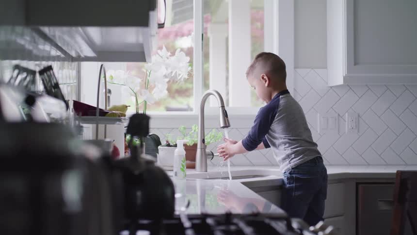 Young boy stands on chair and rubs his hands together under the running water of the kitchen faucet to wash his hands