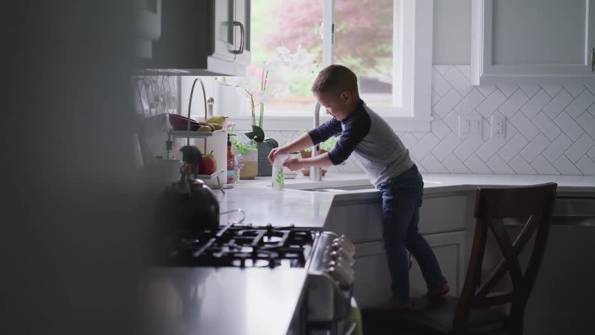 A young boy washes his hands at a kitchen sink while standing on a dining chair