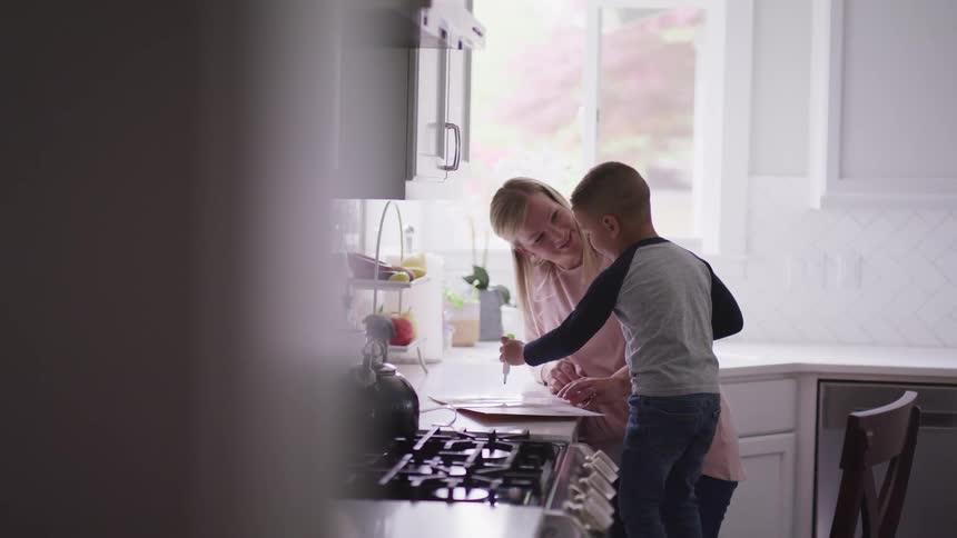 A young boy stands on a chair to reach a kitchen counter with a woman standing right next to him