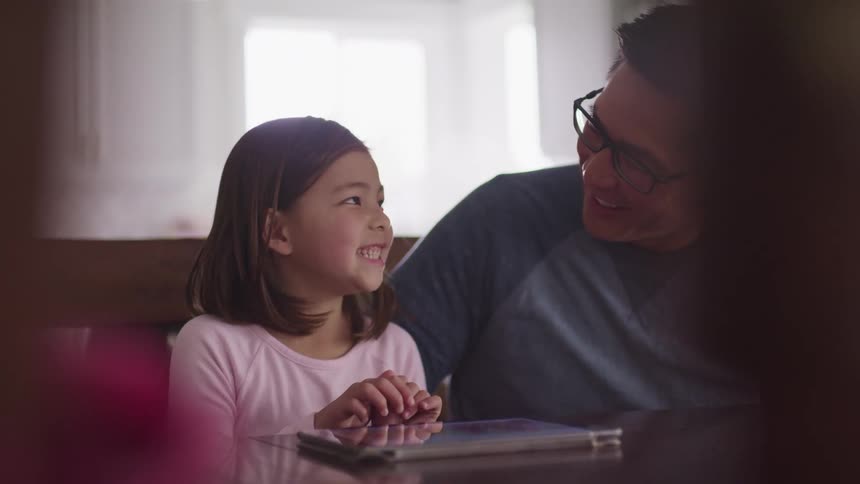Asian daughter with a long sleeve pink t shirt sitting at a table uses a tablet and father wearing glasses looks at her affectionately and laughs