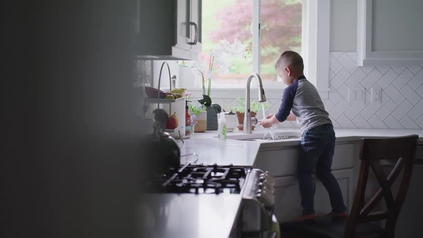 A little boy stands on a chair at the kitchen sink and washes his hands