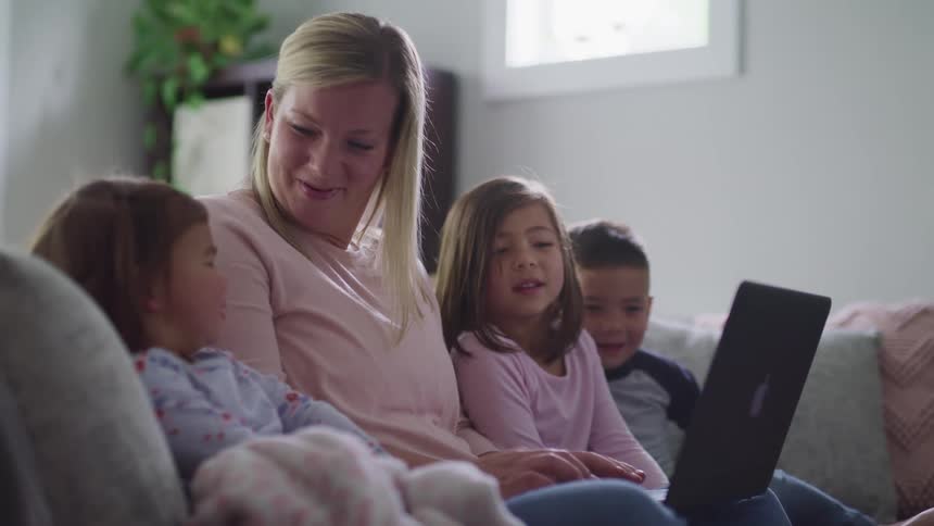 A woman sits on a sofa along with three children and speaks with them while showing them the screen of her laptop