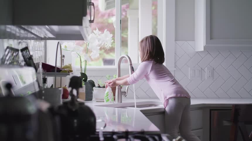 A young girl standing on a stool reaches out to get soap while washing her hands at the kitchen sink