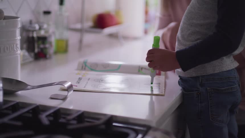 A child draws in a magazine using a green marker as an adult sitting nearby watches