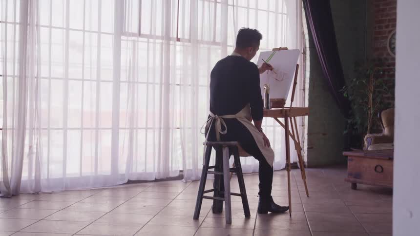 A man paints on an easel set up in an apartment with brick walls and a large window