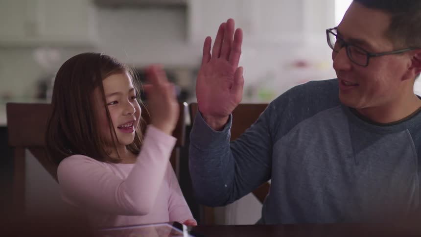 Father and daughter sitting at dining table both raise their hand and give one another a high five and then daughter reaches for the tablet on the table