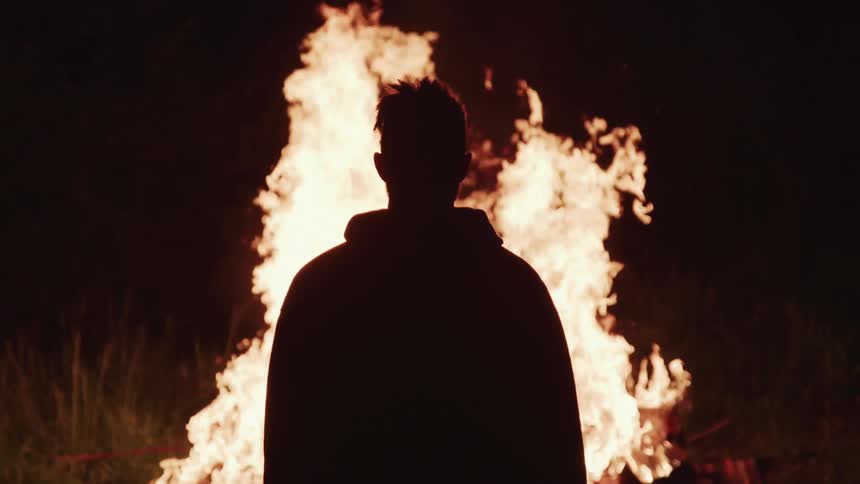 Man with hoodie is silhouetted against a fire that he watches