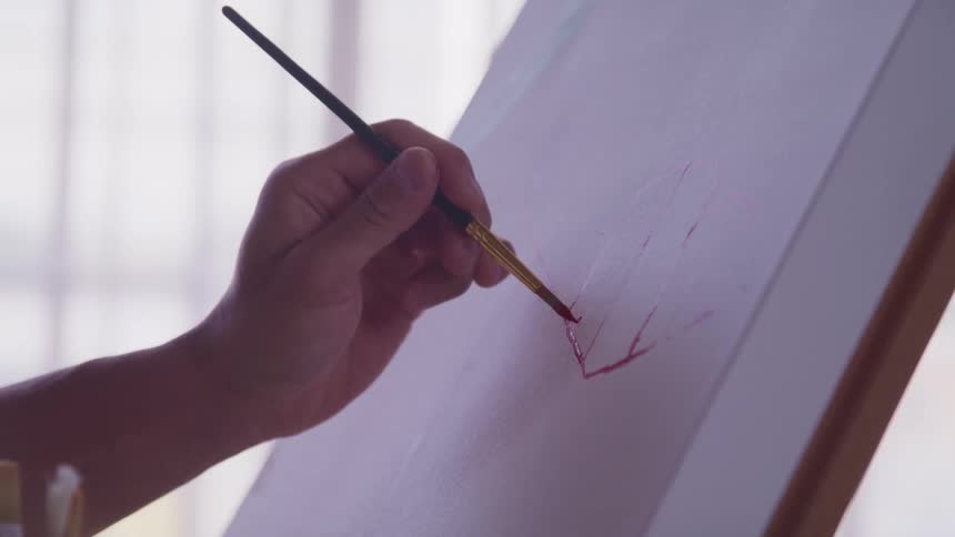 A person uses a thin paint brush to paint on a sheet of paper on a tripod
