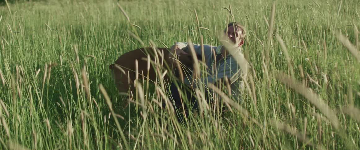 Middle aged man wearing a grey sweater sits in tall grass and strokes a dog