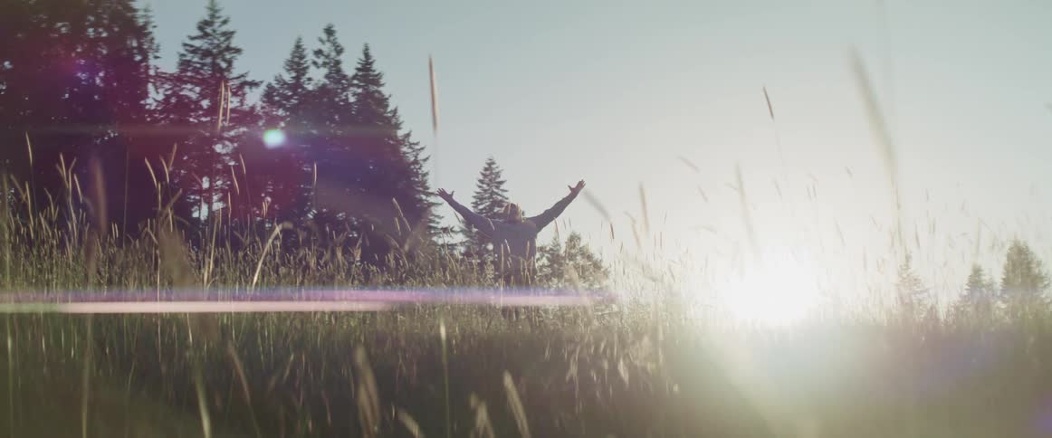 Man stands near pine trees in a grass field with arms wide open at sunrise