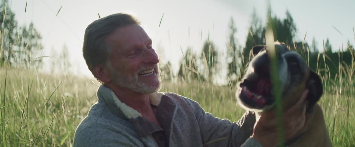 Middle aged man sitting in tall grass pets a dog as the sun shines brightly in a cloudless sky