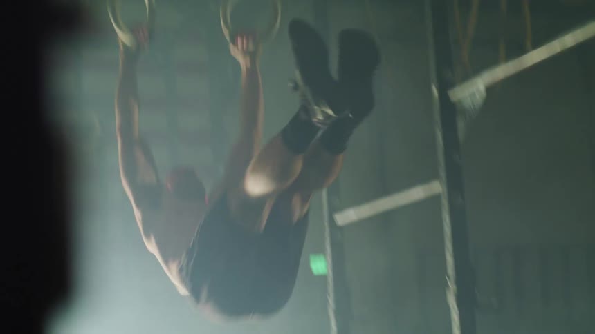 Strong muscular man trains energetically on gymnastic rings in a dimly lit gym