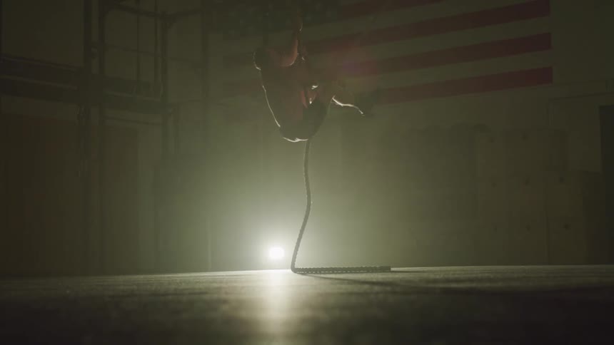A man uses his arms to climb a rope in a dimly lit room