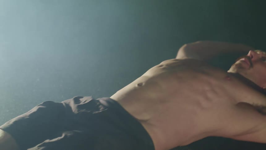 A shirtless man doing crunches in a dark misty room illuminated by a single light