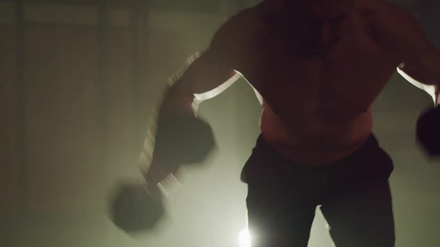 A shirtless man exercises with dumbbells in a dark room illuminated by a single spotlight