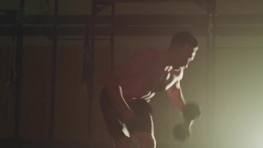 Shirtless strong muscular man exercises with dumbbells on a dimly lit gymnasium