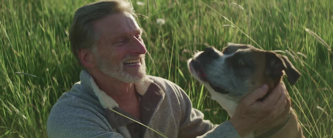 A man is petting his dog outdoors in a grassland area