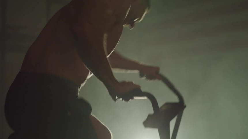 Bare chested man exercises using stationary bike in dark foggy room