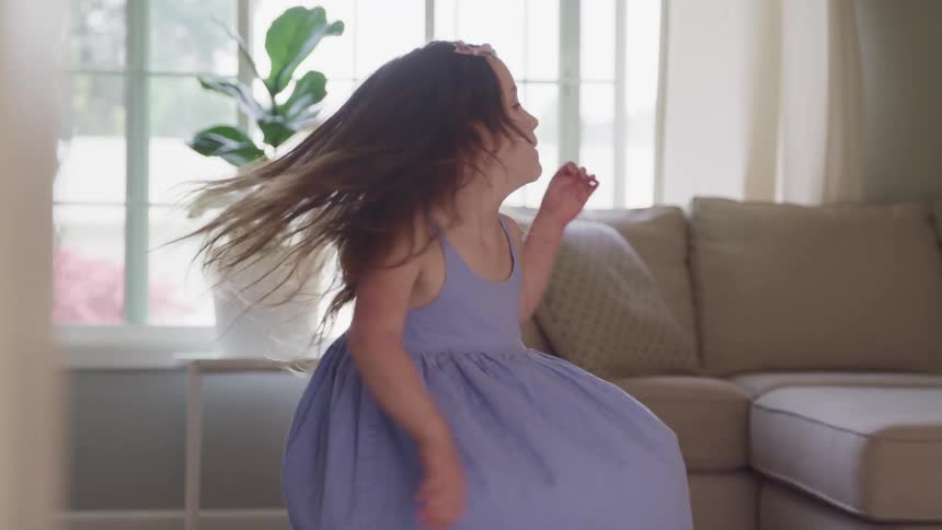 Little child with a blue dress turns around and dances in a living room at home