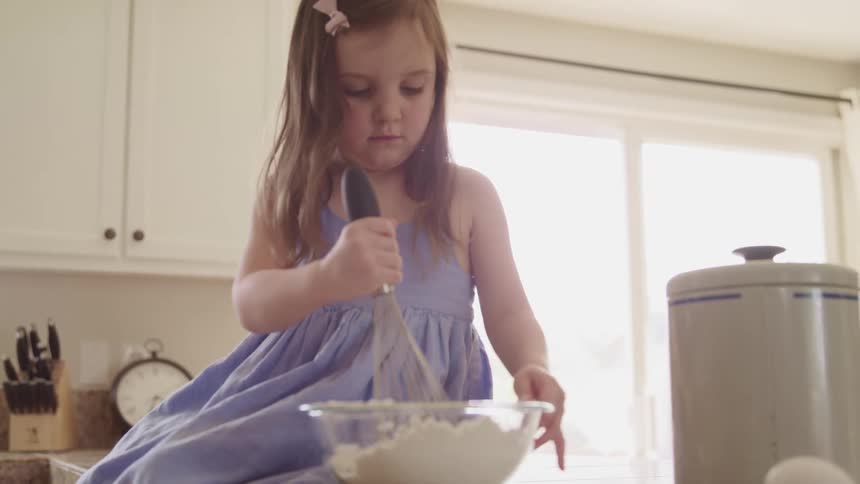 Little girl sits on a kitchen counter and uses a whisk in a glass bowl of flour