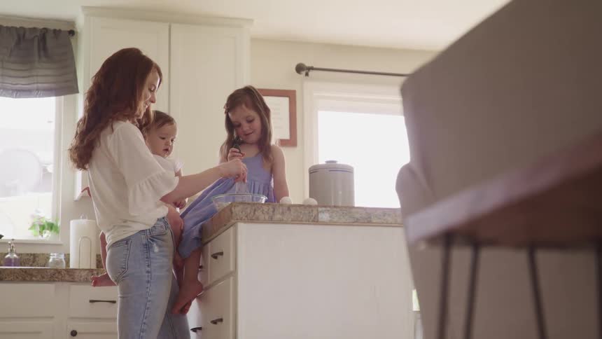A mother holds her baby as she helps an older daughter stir food in a bowl