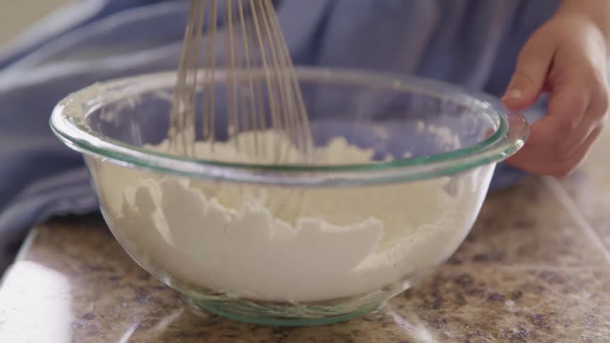 A little girl sitting on a kitchen counter uses a metal whisk to stir powder in a glass bowl