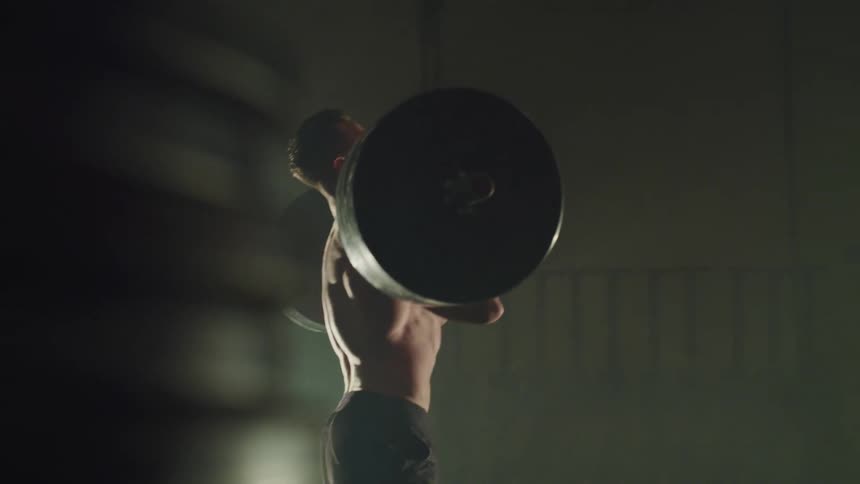 Shirtless strong muscular man deadlifts weights over his head inside a dimly lit gymnasium