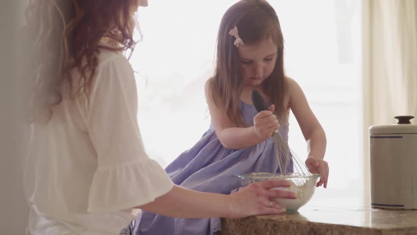 A young girl sits on a kitchen counter in a purple dress and helps her mother stir flour in a mixing bowl