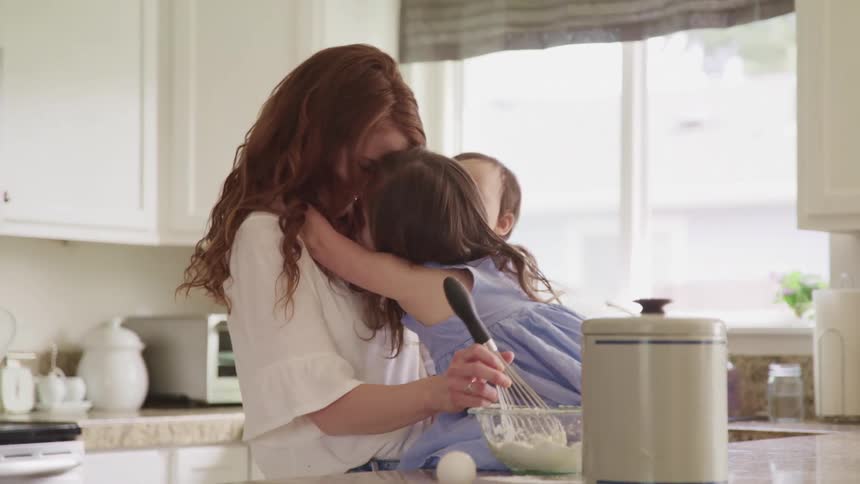 A woman a little girl and a baby in the kitchen as the woman mix flour in a bowl