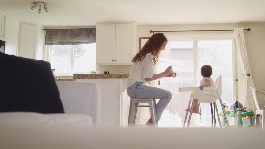 A woman sitting on a stool uses a spoon to feed baby food to a baby in a feeding chair