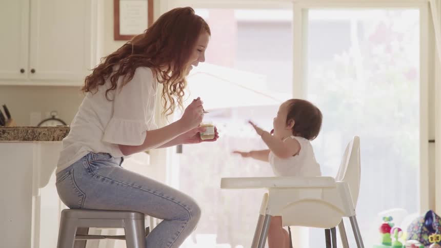 A mother sits on a kitchen stool feeding a baby in a highchair