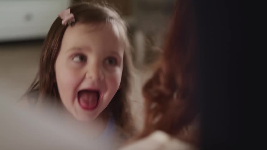 A young girl makes faces at her mother and laughs as they sit on the floor in a bedroom