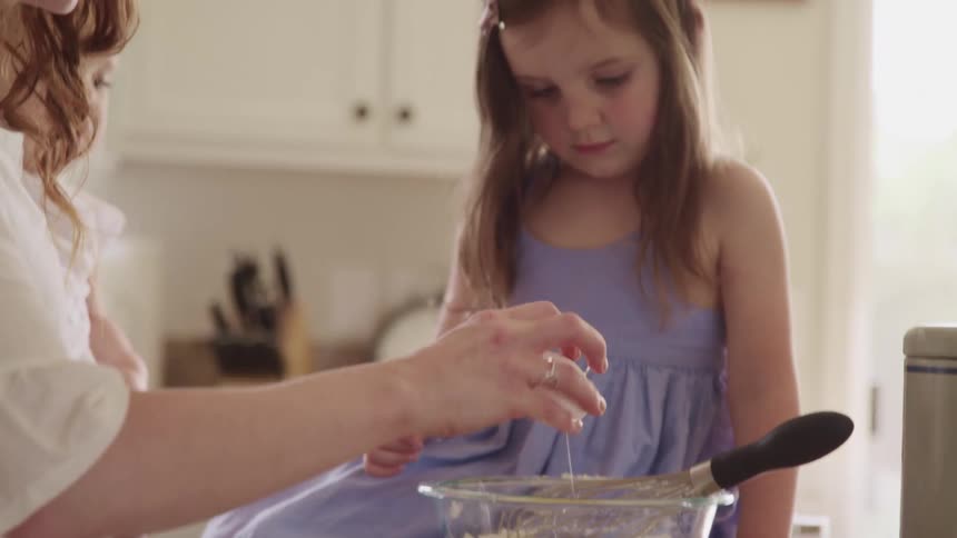 Daughter picks up whisk and starts to stir the ingredients together while sitting on the kitchen counter after mother cracks egg into glass bowl containing flour with one hand