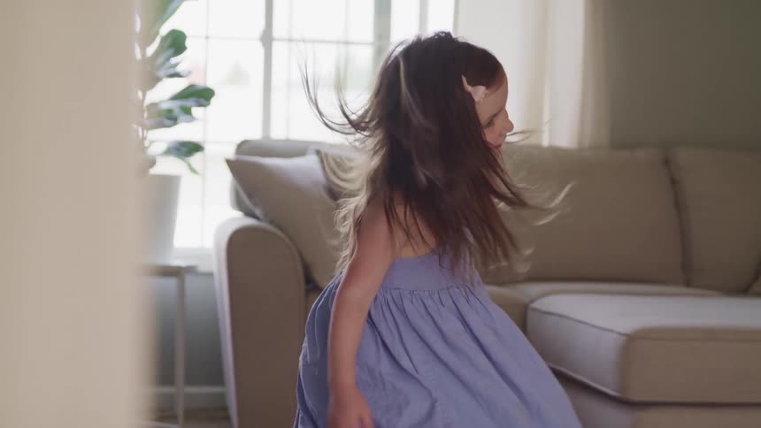 A young girl in a blue dress spins and smiles in a living room