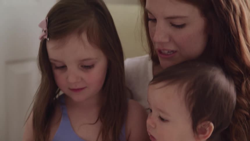 A mother sits at home with both her daughters in her lap as they look at a book