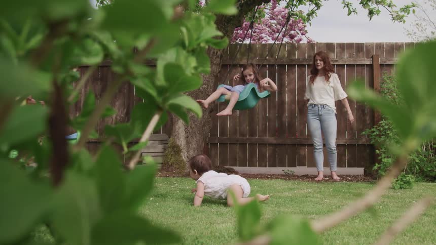 A woman pushes her daughter on a swing in their backyard as her crawls by