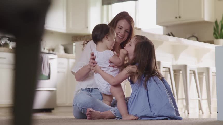 Mother with curly brown hair and a white shirt sits on the ground and plays with her baby boy and little daughter in the kitchen at home