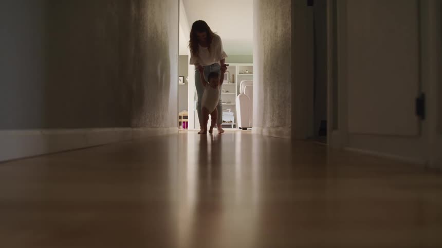 A woman helps a baby to walk down a hallway in a house