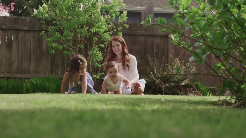Female toddler mimics female baby crawling off the blanket in the back yard while their mother sits on the blanket smiling and laughing with enjoyment