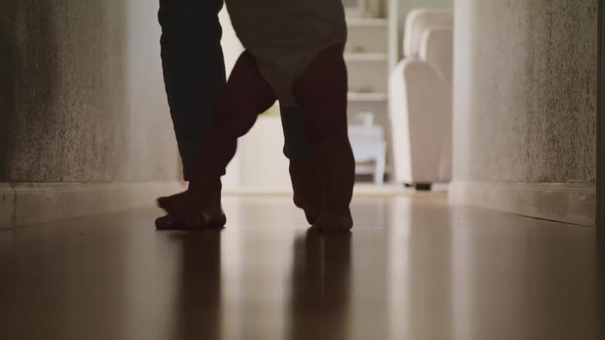 Barefoot mother teaches a baby to walk on a dimly lit hallway at home