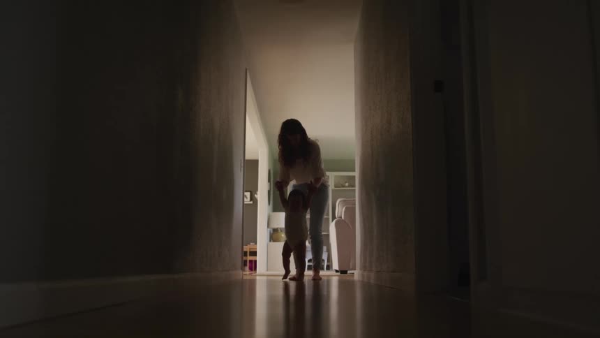 A woman helps a baby to walk down a hallway in a house