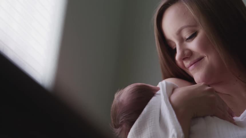 Mother with a long brown hair holds her baby and looks at it affectionately