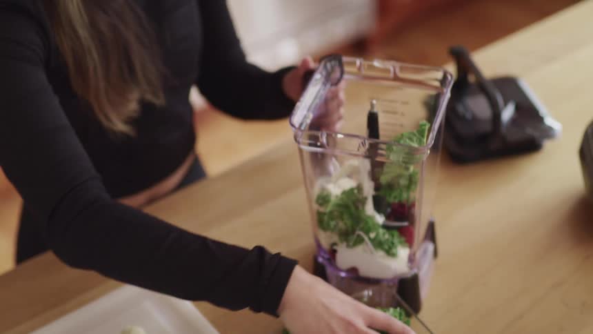 A woman puts vegetables into a blender filled with berries and cream