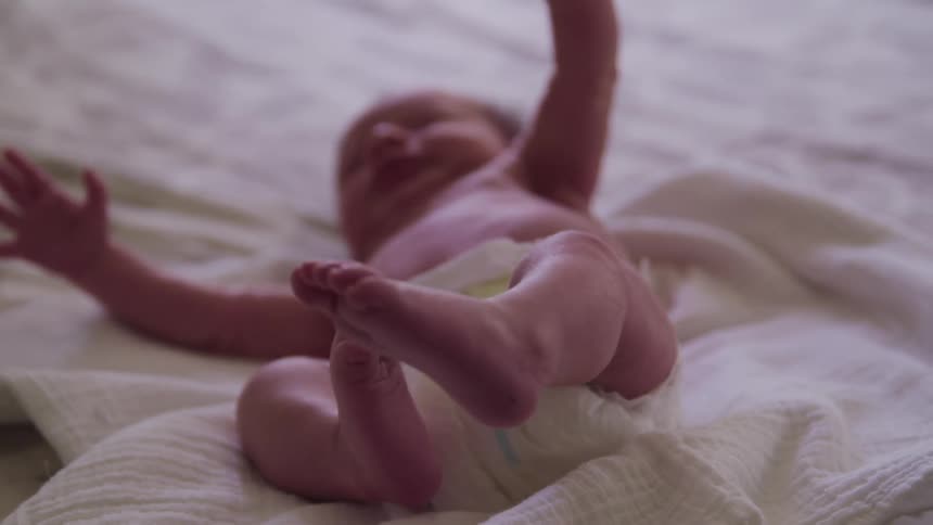 Baby wearing diaper kicks feet into air while lying down on white bed spread