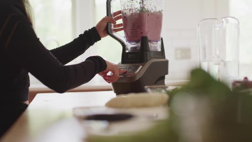 Woman uses blender to mix a juice with fruits and vegetables in the kitchen at home
